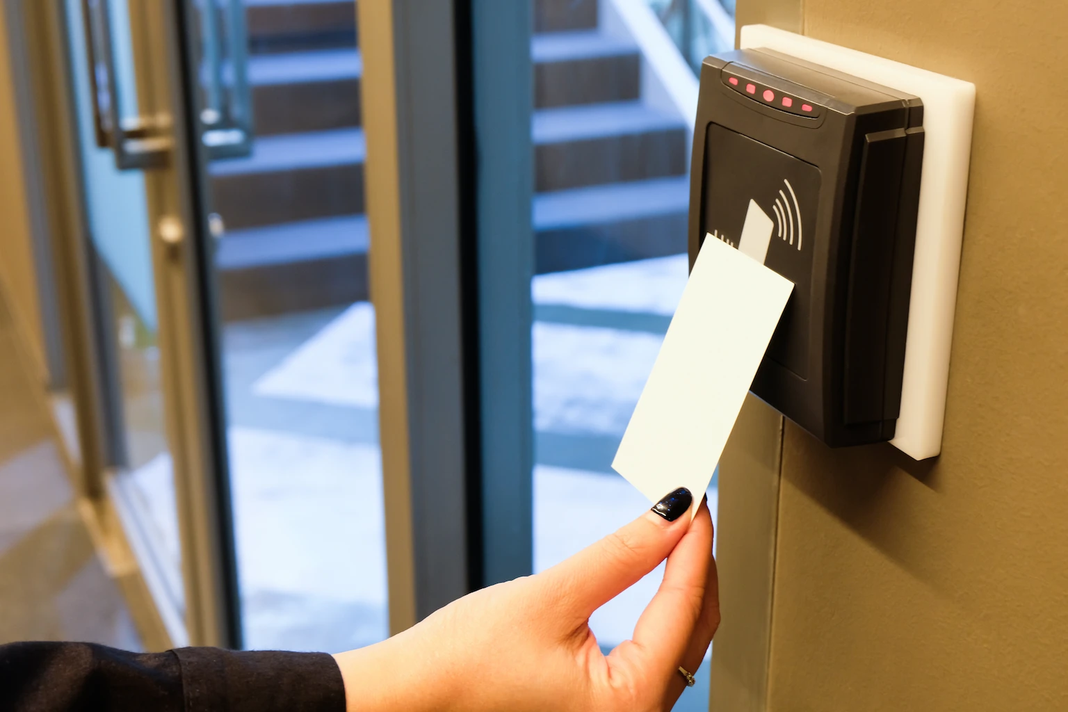 Hand holding an RFID access card near a wall-mounted card reader at a secure glass entry door for controlled building access